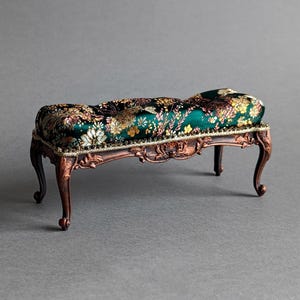 May include: A miniature bench with a dark green floral patterned cushion and ornate bronze-colored legs. The cushion features gold and pink floral designs. The legs have a curved, detailed design, adding to the bench's antique aesthetic.