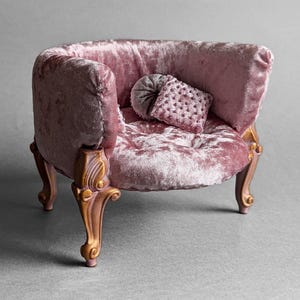May include: A miniature pink velvet armchair with ornate wooden legs and a matching pillow. The chair has a rounded back and a tufted seat. The legs are a light brown colour.