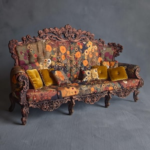 Könnte beinhalten: Ein Miniatursofa mit einem dunkel bronzefarbenen, geschnitzten Holzrahmen mit aufwendigen Verzierungen. Der Bezug zeigt ein mehrfarbiges Blumenmuster in Orange-, Braun- und Goldtönen, akzentuiert durch mehrere kleine, einfarbige senfgelbe Kissen.