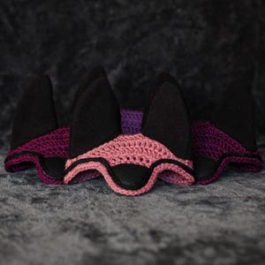 K&ouml;nnte beinhalten: Drei Ohrenhauben mit schwarzen Ohrenabdeckungen. Die Hauben haben ein rosa und lila geh&auml;keltes Design mit schwarzen Lederakzenten. Der Hintergrund ist eine dunkle, strukturierte Oberfl&auml;che.