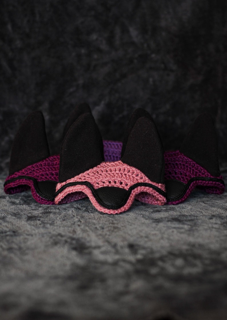 K&ouml;nnte beinhalten: Drei Ohrenhauben mit schwarzen Ohrenabdeckungen. Die Hauben haben ein rosa und lila geh&auml;keltes Design mit schwarzen Lederakzenten. Der Hintergrund ist eine dunkle, strukturierte Oberfl&auml;che.
