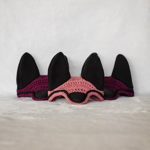 K&ouml;nnte beinhalten: Drei Fliegenohren f&uuml;r Pferde mit schwarzen Ohrensch&uuml;tzern und geh&auml;kelten Stirnb&auml;ndern. Eine Ohrenhaube hat ein rosa Stirnband, die anderen beiden ein dunkelviolettes Stirnband. Die Ohrenhauben werden auf einer hellgrauen Oberfl&auml;che pr&auml;sentiert.