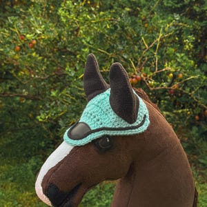 K&ouml;nnte beinhalten: Ein brauner Steckenpferdkopf mit einem wei&szlig;en Streifen auf der Nase und einer t&uuml;rkis geh&auml;kelten Haube. Die Haube hat eine schwarze Bord&uuml;re und schwarze Ohrensch&uuml;tzer. Der Hintergrund ist eine verschwommene Ansicht von gr&uuml;nem Laub.