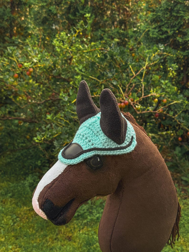 K&ouml;nnte beinhalten: Ein brauner Steckenpferdkopf mit einem wei&szlig;en Streifen auf der Nase und einer t&uuml;rkis geh&auml;kelten Haube. Die Haube hat eine schwarze Bord&uuml;re und schwarze Ohrensch&uuml;tzer. Der Hintergrund ist eine verschwommene Ansicht von gr&uuml;nem Laub.