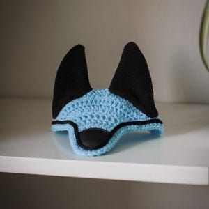 K&ouml;nnte beinhalten: Eine blaue und schwarze H&auml;kel-Pferdehaube. Die Haube hat einen hellblauen H&auml;kelk&ouml;rper mit schwarzen Ohrensch&uuml;tzern und einem Nasenst&uuml;ck aus schwarzem Kunstleder. Sie sch&uuml;tzt die Ohren des Pferdes vor Insekten und L&auml;rm.