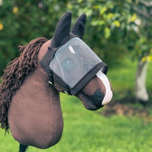 Op de afbeelding: Een bruin stokpaard met manen van donkerbruin garen, een witte bles op de neus en een zwart vliegenmasker van gaas over de ogen en oren. De paardenkop is bevestigd aan een houten stok en is voorzien van zwarte fleece oorbeschermers en randen.