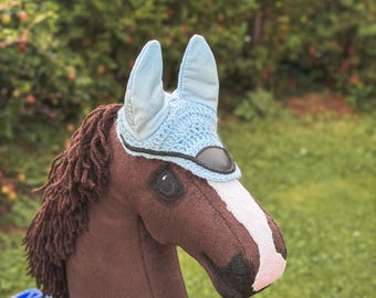 May include: A brown stick horse with a light blue bonnet and a white stripe down its face. The horse has a brown mane and is sitting on a blue plastic crate. The background is a blurred green.