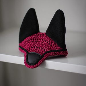 K&ouml;nnte beinhalten: Eine magentafarbene H&auml;kel-Pferdehaube mit schwarzen Ohrensch&uuml;tzern und einem schwarzen Kunstlederakzent. Die Haube soll die Ohren des Pferdes vor Insekten sch&uuml;tzen und L&auml;rm reduzieren.