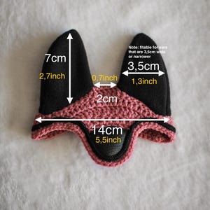 K&ouml;nnte beinhalten: Eine schwarze und rosa geh&auml;kelte Ohrenhaube f&uuml;r ein Pferd, mit den Ma&szlig;en 7 cm hoch, 14 cm breit und Ohren&ouml;ffnungen von 3,5 cm breit. Der Text gibt an, dass sie f&uuml;r Ohren von 3,5 cm Breite oder weniger geeignet ist.
