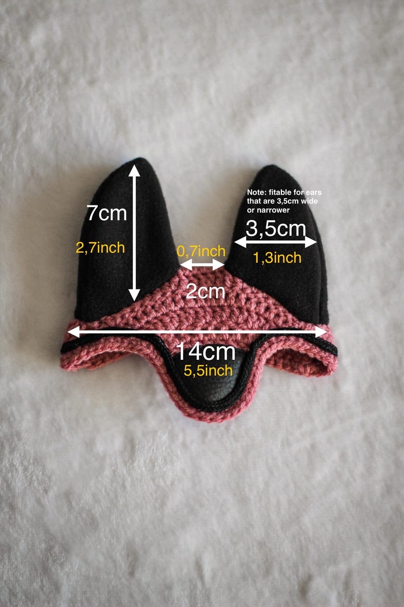 K&ouml;nnte beinhalten: Eine schwarze und rosa geh&auml;kelte Ohrenhaube f&uuml;r ein Pferd, mit den Ma&szlig;en 7 cm hoch, 14 cm breit und Ohren&ouml;ffnungen von 3,5 cm breit. Der Text gibt an, dass sie f&uuml;r Ohren von 3,5 cm Breite oder weniger geeignet ist.