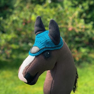 K&ouml;nnte beinhalten: Ein braunes Steckenpferd mit einer blau-schwarzen Reitkappe. Das Pferd hat einen wei&szlig;en Streifen im Gesicht und eine schwarze M&auml;hne. Der Hintergrund ist verschwommenes gr&uuml;nes Laub und Gras.
