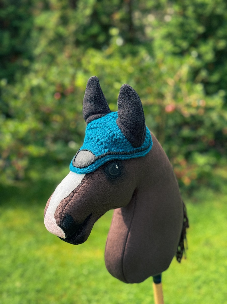 K&ouml;nnte beinhalten: Ein braunes Steckenpferd mit einer blau-schwarzen Reitkappe. Das Pferd hat einen wei&szlig;en Streifen im Gesicht und eine schwarze M&auml;hne. Der Hintergrund ist verschwommenes gr&uuml;nes Laub und Gras.
