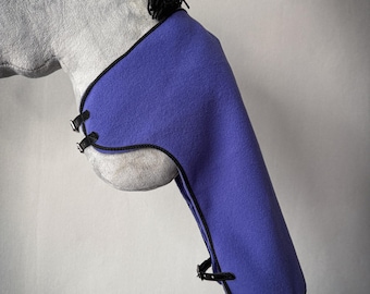 May include: A hobby horse with a light gray plush head and black yarn mane, wearing a purple fleece neck cover. The neck cover features black trim and three black adjustable straps with silver-toned metal buckles, secured around the neck and chest.