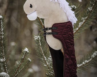May include: A plush white horse head with a burgundy quilted saddle and black straps. The horse is mounted on a wooden stick and is surrounded by a snowy pine tree.