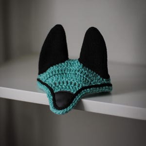 K&ouml;nnte beinhalten: Ein geh&auml;keltes Fuchsohren-Bonnet in Teal und Schwarz. Das Bonnet hat eine geh&auml;kelte Basis in Teal mit schwarzen Ohren und einer Nase aus schwarzem Kunstleder. Der Artikel ist zum Tragen auf dem Kopf bestimmt und wird auf einem wei&szlig;en Regal ausgestellt.
