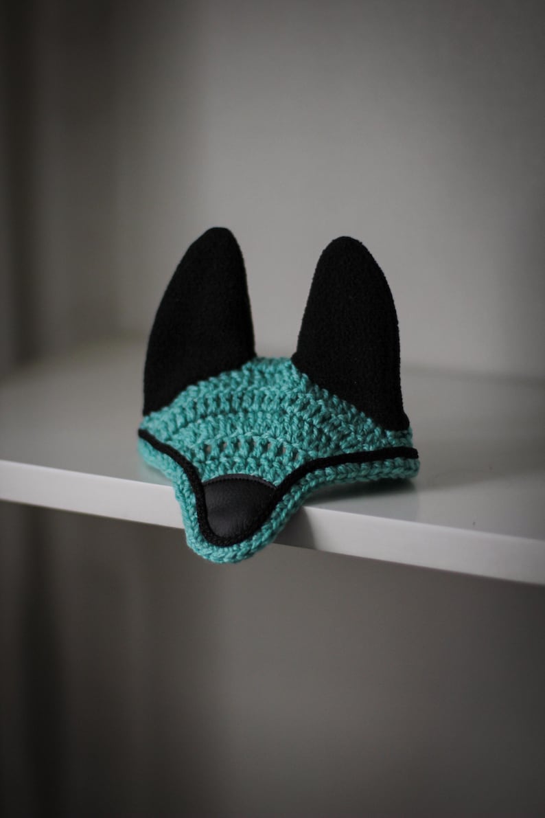 K&ouml;nnte beinhalten: Ein geh&auml;keltes Fuchsohren-Bonnet in Teal und Schwarz. Das Bonnet hat eine geh&auml;kelte Basis in Teal mit schwarzen Ohren und einer Nase aus schwarzem Kunstleder. Der Artikel ist zum Tragen auf dem Kopf bestimmt und wird auf einem wei&szlig;en Regal ausgestellt.