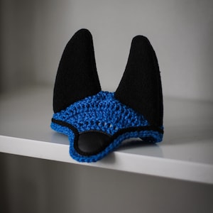 K&ouml;nnte beinhalten: Eine handgefertigte Fliegenhaube f&uuml;r Pferde in Schwarz und Blau. Die Haube hat zwei aufrechte schwarze Ohrensch&uuml;tzer und ein blau geh&auml;keltes Kopfst&uuml;ck mit schwarzem Besatz. Der Artikel dient dazu, die Ohren eines Pferdes vor Insekten zu sch&uuml;tzen.