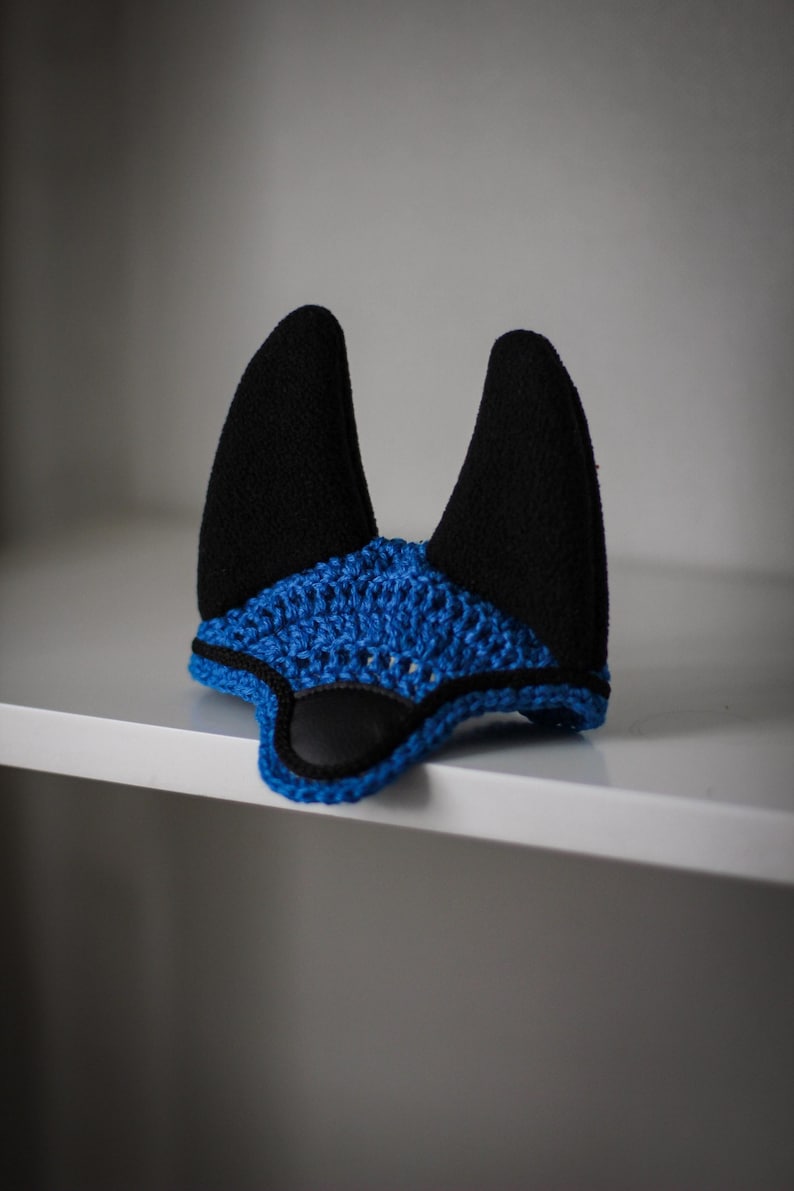 K&ouml;nnte beinhalten: Eine handgefertigte Fliegenhaube f&uuml;r Pferde in Schwarz und Blau. Die Haube hat zwei aufrechte schwarze Ohrensch&uuml;tzer und ein blau geh&auml;keltes Kopfst&uuml;ck mit schwarzem Besatz. Der Artikel dient dazu, die Ohren eines Pferdes vor Insekten zu sch&uuml;tzen.