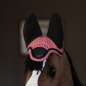 K&ouml;nnte beinhalten: Ein brauner Steckenpferdkopf mit einem rosa geh&auml;kelten Stirnband und schwarzen Ohrensch&uuml;tzern. Das Pferd hat einen wei&szlig;en Bl&auml;sse und eine schwarze M&auml;hne. Das Stirnband hat einen schwarzen Leder-Augenschutz.