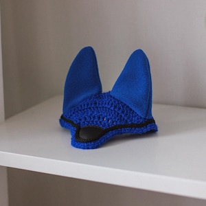 Può includere: Un cappuccio blu per cavalli, che combina materiali all'uncinetto e feltro. Ha due copri orecchie verticali e una sezione all'uncinetto per la fronte. Il bordo inferiore è rifinito in nero. Questo articolo è progettato per proteggere le orecchie di un cavallo da insetti e rumori.