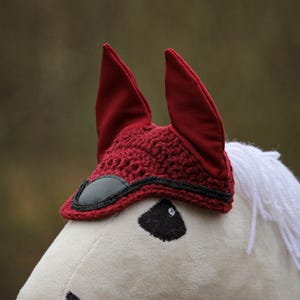 Könnte beinhalten: Ein roter gehäkelter Fliegenhaube für Pferde mit schwarzem Besatz und Ohrenklappen. Die Haube sitzt auf einem weißen Plüschpferdekopf.