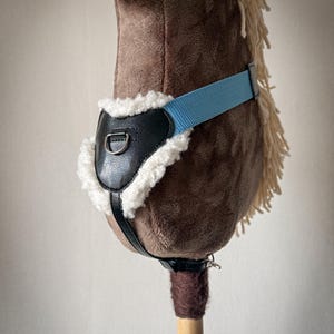 May include: A brown plush hobby horse with a light blue strap and black leather harness. The horse has a cream-coloured mane and a wooden stick handle. The harness is lined with white faux fur.