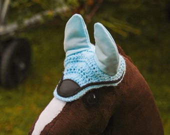 May include: A brown plush horse head with a light blue crocheted bonnet and ear covers. The horse has a white stripe down its face and a pink nose. The background is blurred with green grass and foliage.