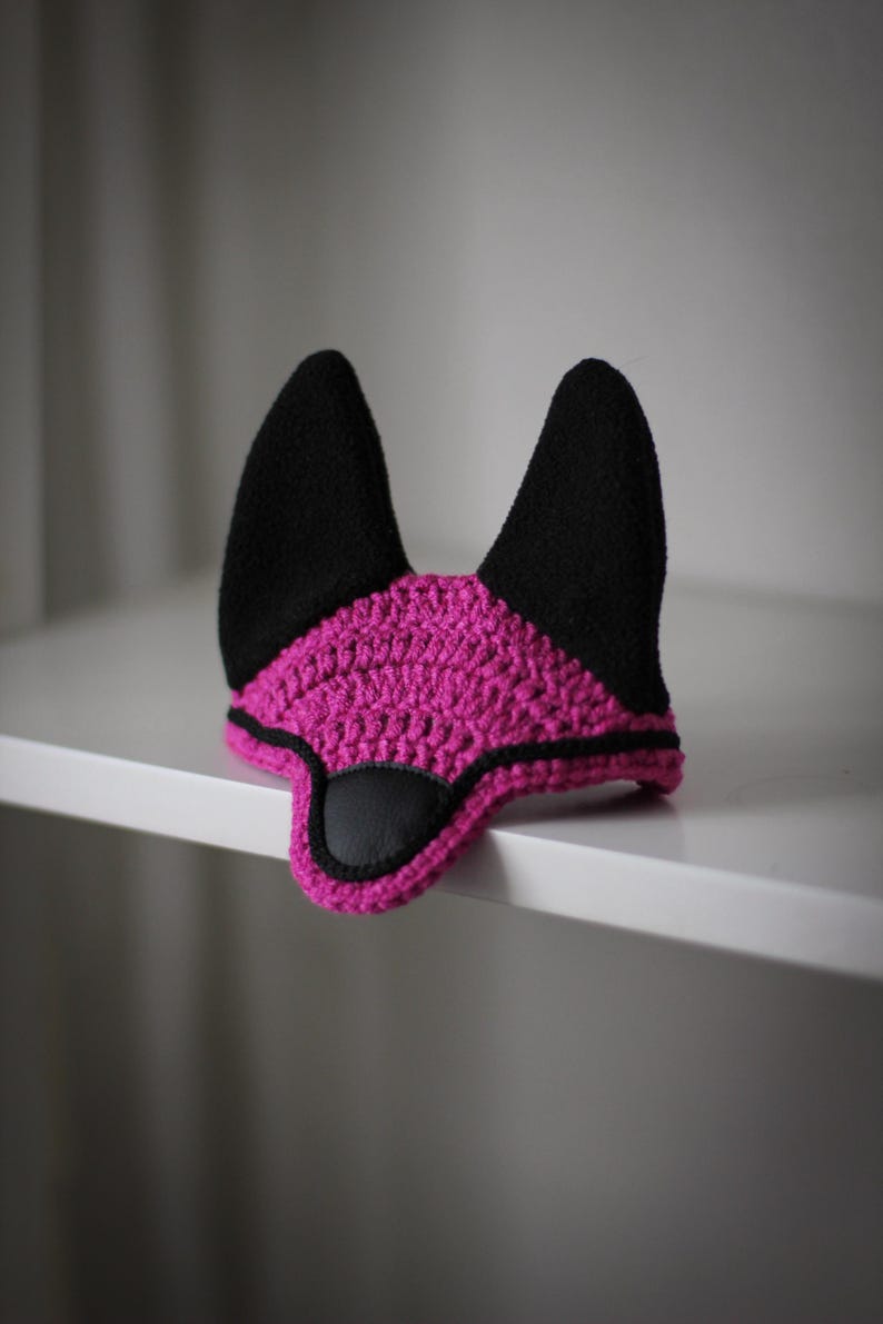 K&ouml;nnte beinhalten: Eine handgefertigte H&auml;kelpferdehaube in leuchtendem Pink mit schwarzen Akzenten. Die Haube hat schwarze, aufrechte Ohren und einen schwarzen Kunstleder-Augenschutz. Die Kanten sind mit schwarzem Garn verziert. Dieses Reitzubeh&ouml;r dient zum Schutz der Ohren eines Pferdes.