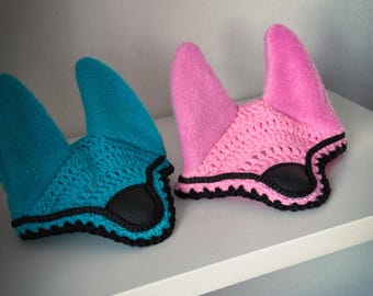 May include: Two handmade horse bonnets. One is turquoise with a black trim and the other is pink with a black trim. Both bonnets have a crochet base and a soft, upright ear covering. The bonnets are displayed on a white shelf.