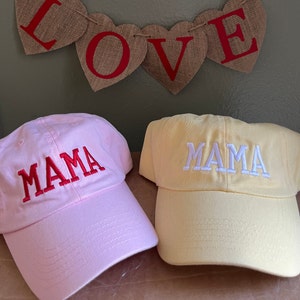 May include: Two baseball caps with the word "MAMA" embroidered on them. One cap is pink with red embroidery and the other is yellow with white embroidery.