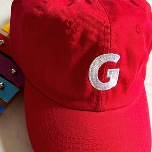 May include: A bright red baseball cap with a curved brim. The cap features a white embroidered "G" on the front. The hat is made of a soft fabric and has a classic, casual style.