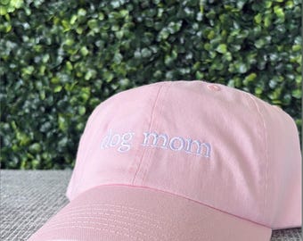 May include: A light pink baseball cap with the embroidered text "dog mom" in white. The cap has a curved brim and a structured crown. The background is a green hedge.
