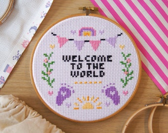 May include: A purple and white embroidery hoop with the text "Welcome to the World" surrounded by a rainbow, flowers, and baby footprints.