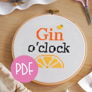 Könnte beinhalten: Ein weißer Stickrahmen mit dem Text "Gin o'clock" in schwarzen und orangen Buchstaben. Unter dem Text befindet sich eine gestickte Zitronenscheibe in Gelb und Orange. Der Rahmen liegt auf einer Holzoberfläche.