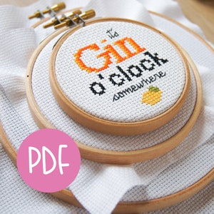 Könnte beinhalten: Ein weißer Stickrahmen mit dem Text "It's Gin o'clock somewhere" in schwarzem und orangefarbenem Garn. Der Rahmen liegt auf einem weißen Stoffuntergrund.