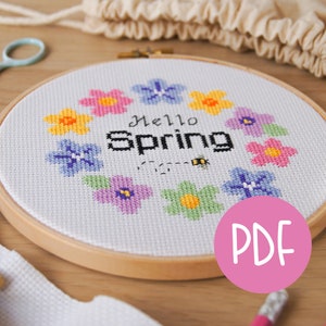 Puede incluir: Un aro de bordado blanco con un diseño de punto de cruz que presenta una corona de flores coloridas y el texto "Hello Spring". Una abeja está bordada debajo del texto. El aro está sobre una superficie de madera con un círculo rosa con la palabra "PDF" en blanco.