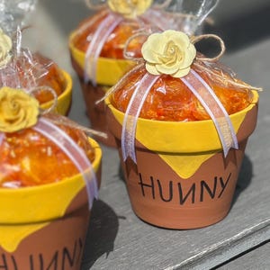 May include: Three small terracotta pots decorated with yellow paint and the word "Hunny" are filled with orange jelly candies and topped with a yellow flower and white ribbon.