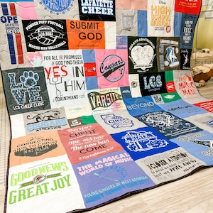 May include: A colorful patchwork quilt made from t-shirts. The quilt features various logos, slogans, and designs, including "Lafayette Christian", "Powder Puff Football", "Submit Yourselves Therefore to God", "Yes In Him", "The Magical World of Music", and "Good News Great Joy".