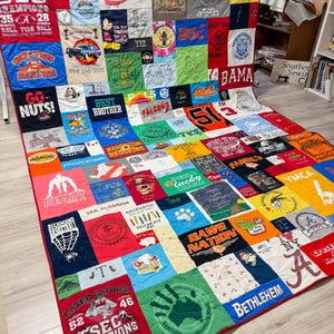 Pode incluir: Um edredom feito à mão composto por várias camisetas coloridas, cada uma com gráficos e texto exclusivos. O edredom tem uma borda vermelha e é exibido em uma moldura branca. As camisetas exibem logotipos, nomes de equipes e outros designs.