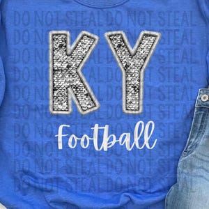 May include: A blue sweatshirt with the letters "KY" in a sparkly design and the word "Football" in white. The phrase "DO NOT STEAL" is printed repeatedly on the shirt. The image also includes a pair of jeans and silver sneakers.