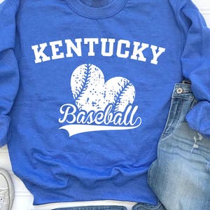 May include: A blue sweatshirt with the text "Kentucky Baseball" and baseball graphics. The sweatshirt is paired with blue jeans and silver sneakers. The items are arranged on a white surface.