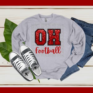 Może przedstawiać: Szara bluza z nadrukiem czerwonego brokatu &bdquo;OH&rdquo; i napisem &bdquo;football&rdquo; z przodu. Bluza leży na drewnianej powierzchni z parą czarnych but&oacute;w z pł&oacute;tna i parą niebieskich dżinsowych szort&oacute;w.