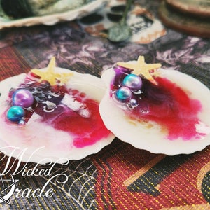 May include: Two white seashell-shaped resin ornaments with a pink and purple swirl design. Each ornament has a gold starfish and iridescent purple and blue beads.