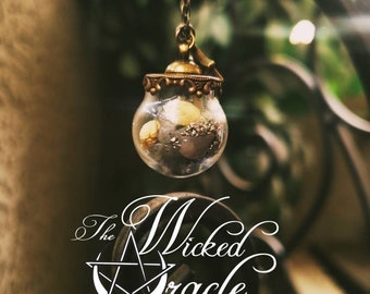 May include: A small, clear glass pendant with a gold-colored metal cap and chain, filled with small, dried flowers and other natural elements. The pendant is hanging in front of a blurred background. The text "The Wicked Oracle" is displayed in white.