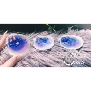 May include: Three white seashells filled with blue and purple resin, gold glitter, and small seashells. The shells are on a fluffy grey surface.
