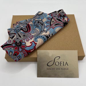 May include: A colorful paisley patterned scarf in shades of blue, red, and gray, resting on a brown box. A small card with the name "Sofia" and the phrase "Macht die Fliege" is in the foreground.