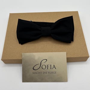 May include: A black bow tie rests on a brown cardboard box. A small card with the name "Sofia" and the phrase "Macht die Fliege" is placed in front of the box. The bow tie is made of a soft, matte material.