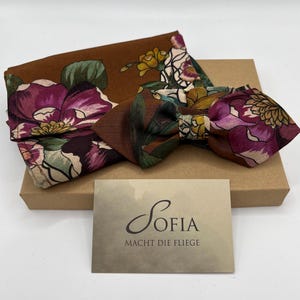 May include: A brown floral patterned bow tie and matching pocket square set. The set features a dark brown background with large purple and white flowers. A business card with the name "Sofia" is also included.