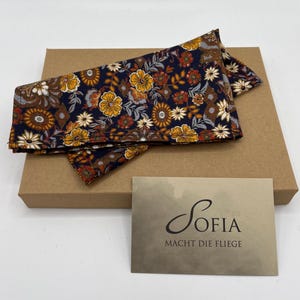 May include: Two floral patterned pocket squares in navy, yellow, and brown, resting on a brown cardboard box. A business card with the name "Sofia" and the phrase "Macht die Fliege" is in the foreground.