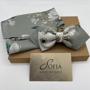 May include: A gray floral bow tie and matching pocket square set. The bow tie is pre-tied and features a white floral pattern. The set is presented on a brown box with a card that reads "Sofia Macht Die Fliege".